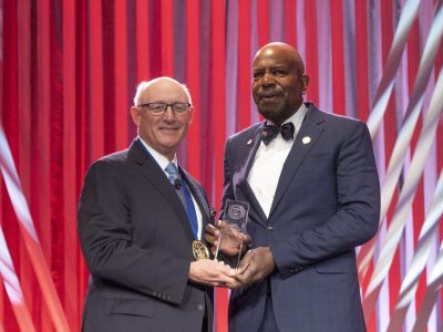 Two men stand on a stage with a red curtain backdrop while holding an award plaque together. One man wears a dark suit and glasses with a medal around his neck, and the other wears a blue suit with a bow tie and lapel pins. Both are smiling during the award presentation.