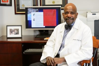 A man wearing a white medical coat and bow tie sits at a desk in an office, smiling toward the camera. Behind him is a computer monitor displaying a book cover titled “Regenerative Engineering,” along with framed certificates and a family photograph on the desk.