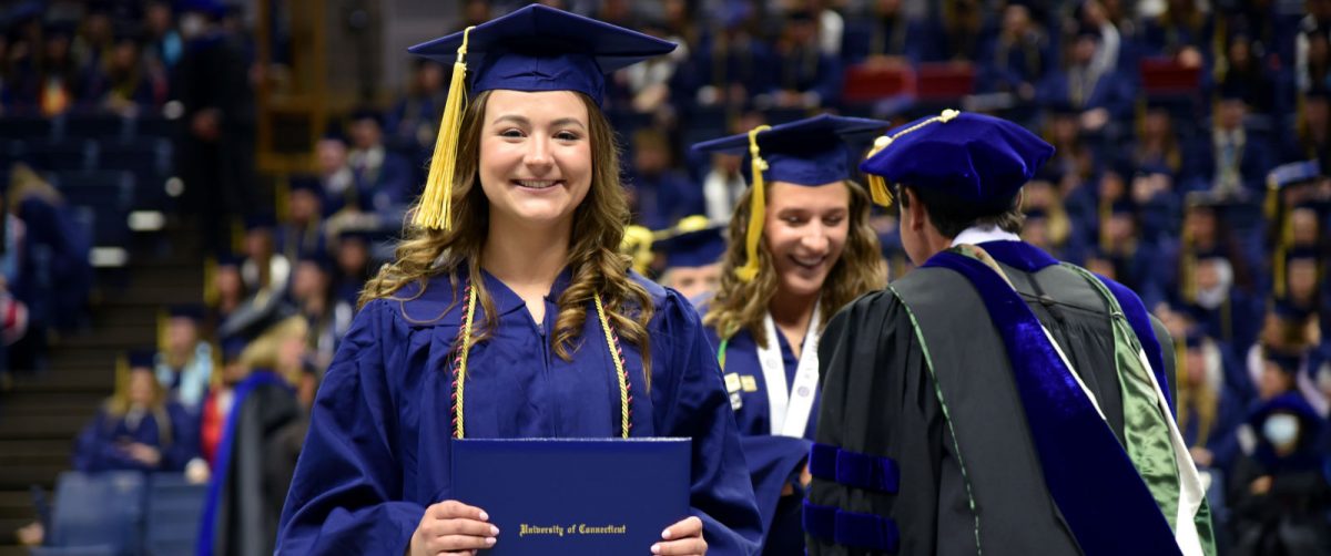 A smiling graduate in a blue cap and gown holds a diploma cover during a commencement ceremony, with other graduates and faculty in academic regalia visible in the background.