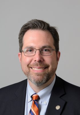 Man with short brown hair, glasses, and a trimmed beard wearing a dark suit and orange striped tie, photographed against a plain gray background.