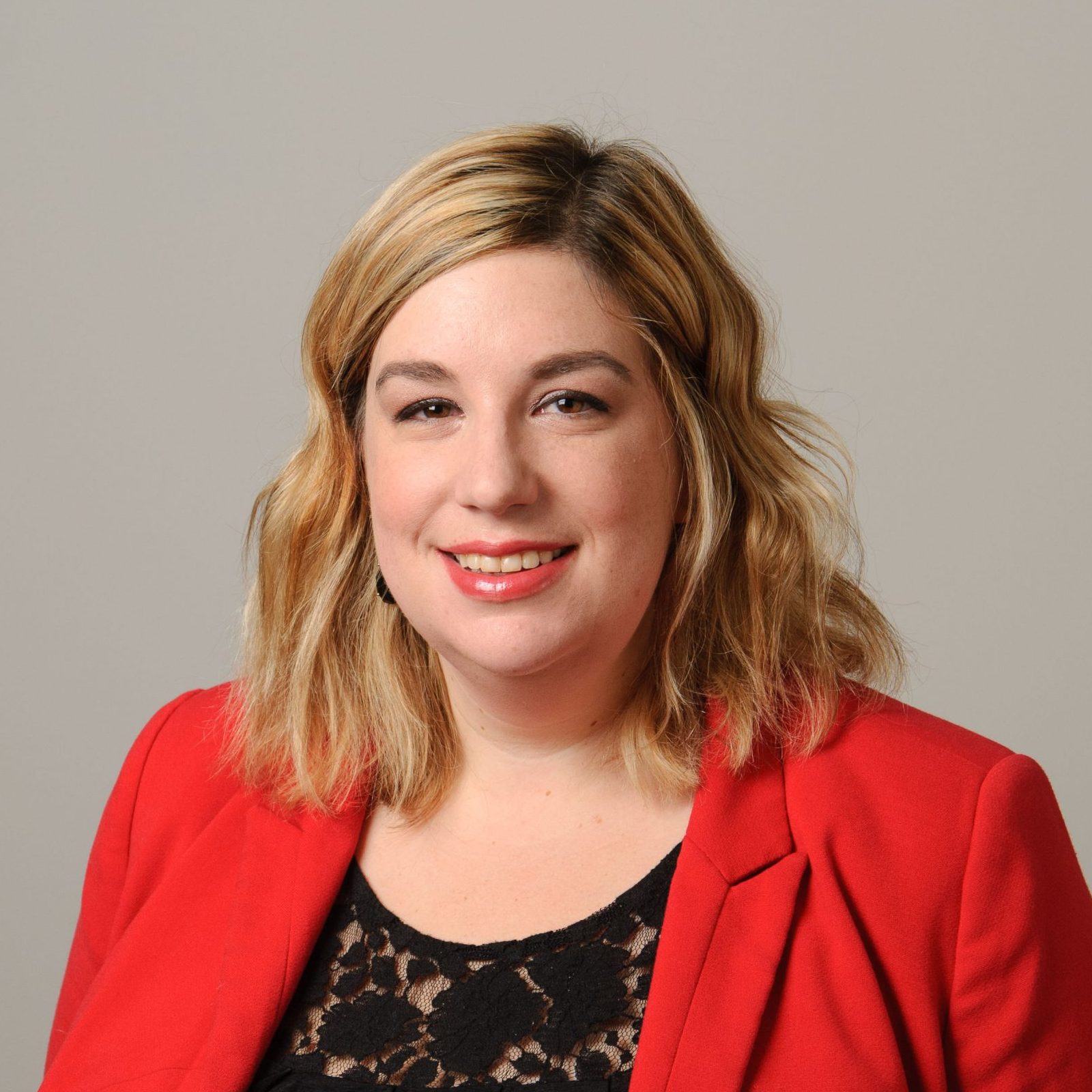 Professional headshot of a smiling woman with shoulder-length blonde hair, wearing a bright red blazer over a black lace top, posed against a neutral gray background.