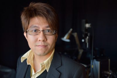 A man wearing glasses and a dark blazer over a light-colored shirt looks at the camera, with a softly blurred indoor background behind him.