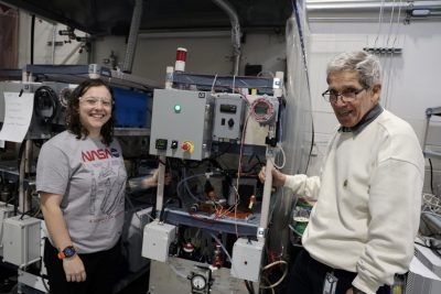 Two people standing beside laboratory equipment, smiling and posing in a research lab setting.