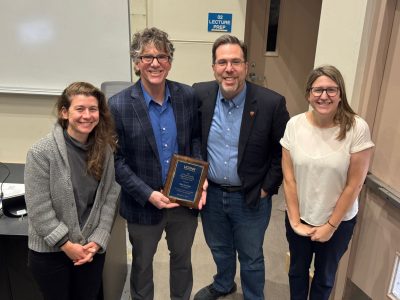 Four colleagues standing together in a classroom, smiling for a group photo. The person second from the left is holding a framed award plaque.