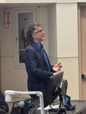 Speaker standing at a lecture podium, gesturing while speaking, with a computer monitor and microphone in front of them in a classroom setting.