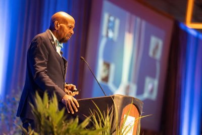 Dr Laurencin speaks at a podium with a microphone, gazing at a projected presentation displaying diagrams. The setting is professional and well-lit.