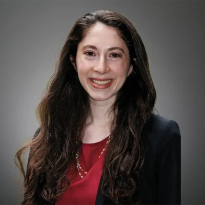 Professional headshot of a woman with long dark brown hair worn down, smiling at the camera. She is wearing a red blouse, a black blazer, and a necklace. The background is a neutral gray studio backdrop.