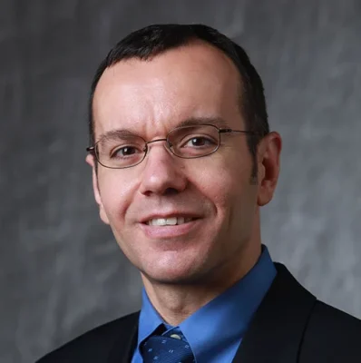 Professional headshot of a man wearing glasses, a dark suit jacket, blue dress shirt, and patterned blue tie. He is smiling slightly and facing the camera against a neutral gray studio background.