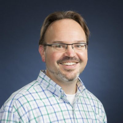 Professional headshot of a man with short light brown hair, glasses, and a trimmed beard and mustache. He is smiling and wearing a light-colored plaid button-down shirt. The background is a dark blue studio backdrop.