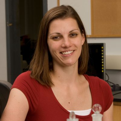 Woman in a red top smiling at a desk, with glass lab equipment in the foreground and an office or lab setting in the background.