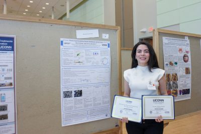 A woman stands beside a research poster at a UConn engineering poster session, holding two certificates, including a People’s Choice Award. Additional posters and display boards are visible in the background of the indoor academic setting.