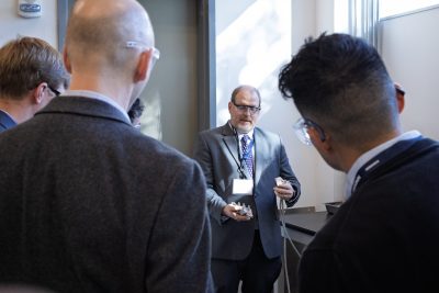 A man in a suit and safety glasses stands near a window, holding small mechanical components while speaking to a small group of people. The audience, also wearing safety glasses, is seen from behind and appears to be listening attentively. The setting resembles a laboratory or demonstration space with a countertop and equipment visible in the background.