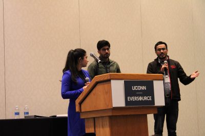 Three people stand at a wooden podium labeled “UCONN EVERSOURCE” during a presentation. A woman in a blue dress holds a microphone and looks toward two men beside her. One man stands behind the podium, and the other, wearing glasses and a dark jacket, speaks into a microphone while gesturing with his hand. A table with water bottles is visible to the side against a plain beige conference room wall.