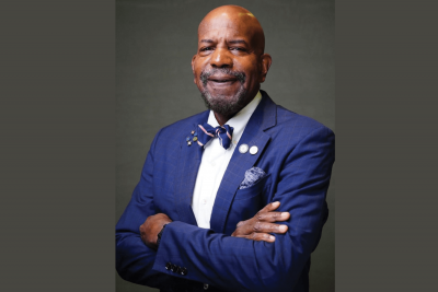 Portrait of a man wearing a blue suit jacket, white shirt, and patterned bow tie, standing with arms crossed against a neutral gray background. He is smiling slightly and wearing several lapel pins and a pocket square.