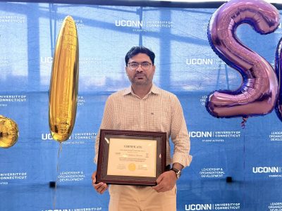 A person stands in front of a UConn Student Activities backdrop holding a framed certificate, with celebratory balloons displayed on either side.