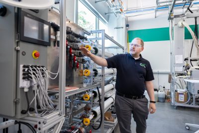A researcher in safety glasses operates and monitors a membrane filtration pilot system with control panels, sensors, and piping inside a chemical engineering laboratory.
