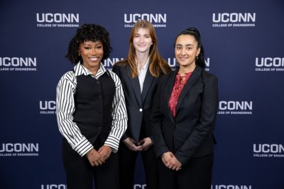 Three students stand together in professional attire in front of a UConn College of Engineering backdrop during Senior Design Demonstration Day, posing for a team photo.