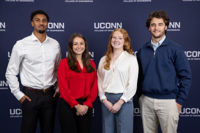Four students pose together in business casual attire in front of a UConn College of Engineering backdrop during Senior Design Demonstration Day.