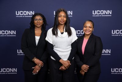 Three students pose together in front of a UConn College of Engineering backdrop during Senior Design Demonstration Day, dressed in professional attire and smiling at the camera.