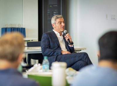 A man in a suit speaks into a microphone while seated at the front of a workshop, with attendees visible in the foreground.