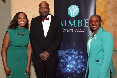Three attendees pose together at an American Institute for Medical and Biological Engineering event in front of an AIMBE banner.